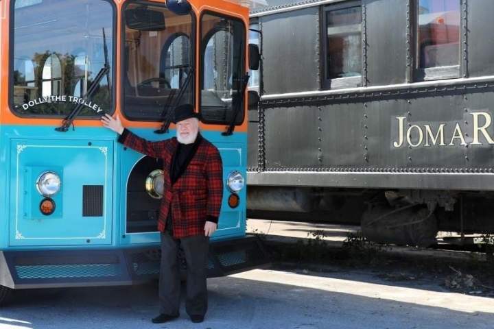 Person in red plaid jacket and hat standing by a blue and orange trolley named 'Dolly the Trolley'.
