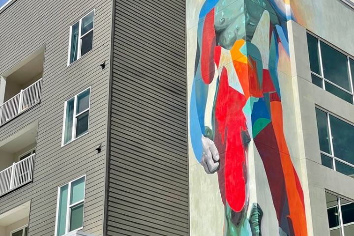 Colorful mural of a statue on a modern building facade against a blue sky.