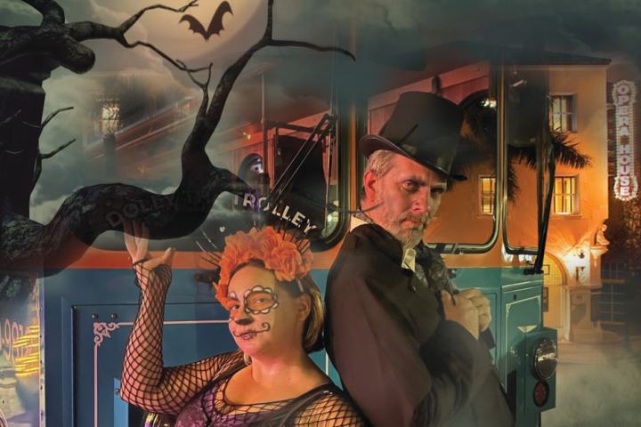 Two people in Halloween costumes by a trolley with a spooky backdrop of a bat and an opera house.