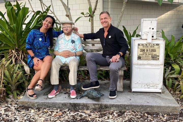 Two people sit smiling on a bench with a statue between them, surrounded by plants and a newspaper dispenser nearby.