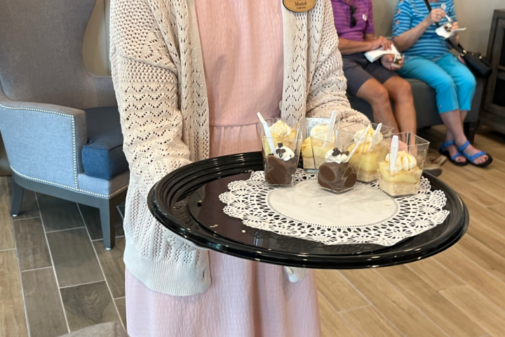 Person holding a tray with desserts in cups; people seated in background.