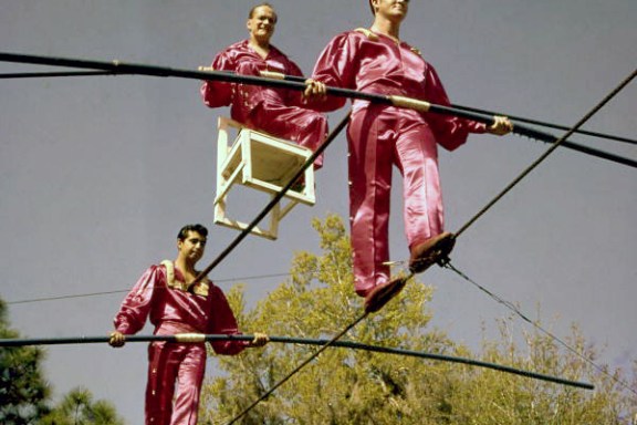Three men in red outfits perform on a high wire, one in a chair.