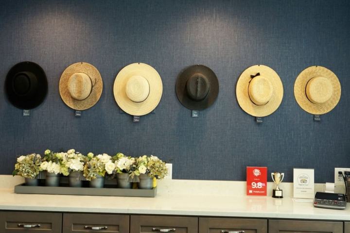 Six straw hats and a row of flowers on a table against a blue wall.