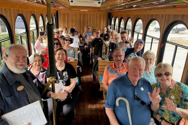 A group of people smiling and waving while seated inside a wooden trolley bus.