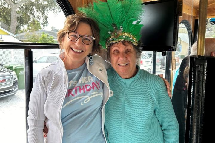 Two women smiling; one wears a green feathered crown inside a wooden interior.