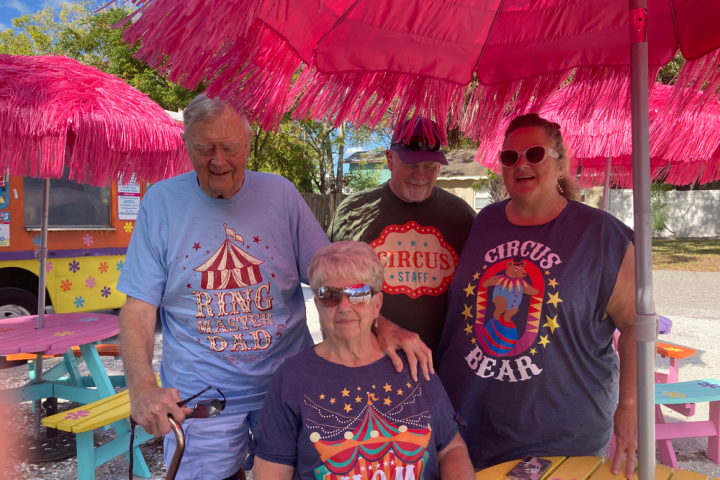 Four people in circus-themed shirts at a colorful outdoor seating area with pink umbrellas.