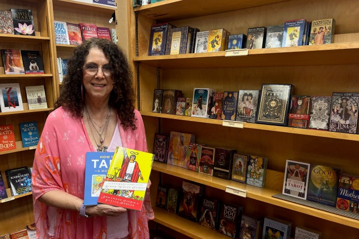 Person in a pink shawl holding tarot books in a bookstore with various tarot decks on shelves.