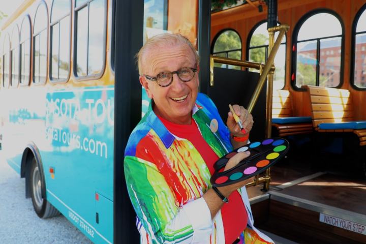 Smiling man in colorful outfit holding paint palette in front of a tour bus.