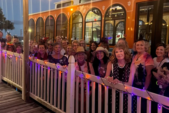 Group of women in colorful hats standing by a lit-up fence and an orange trolley.