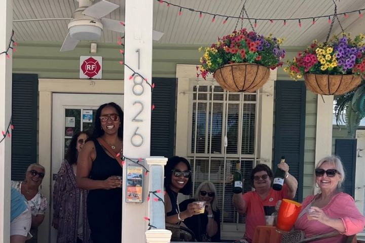 Group of people celebrating on a porch with drinks, flowers, and house number 1826 visible.