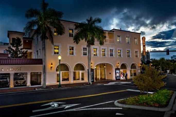 A lit building with arched windows and palm trees at dusk, under a cloudy sky.