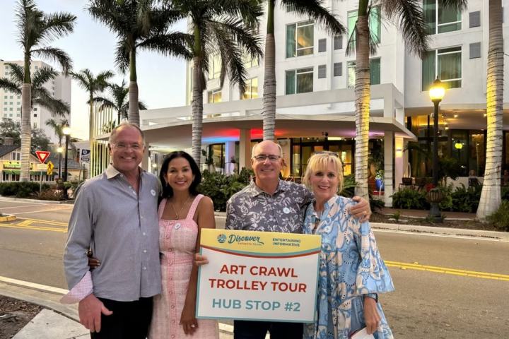 Four people holding 'Art Crawl Trolley Tour Hub Stop #2' sign stand outside a building at sunset.