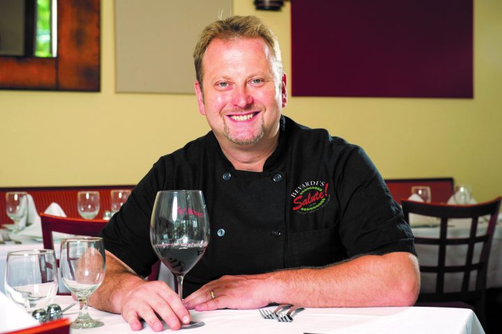 a man sitting at a table with wine glasses and smiling at the camera