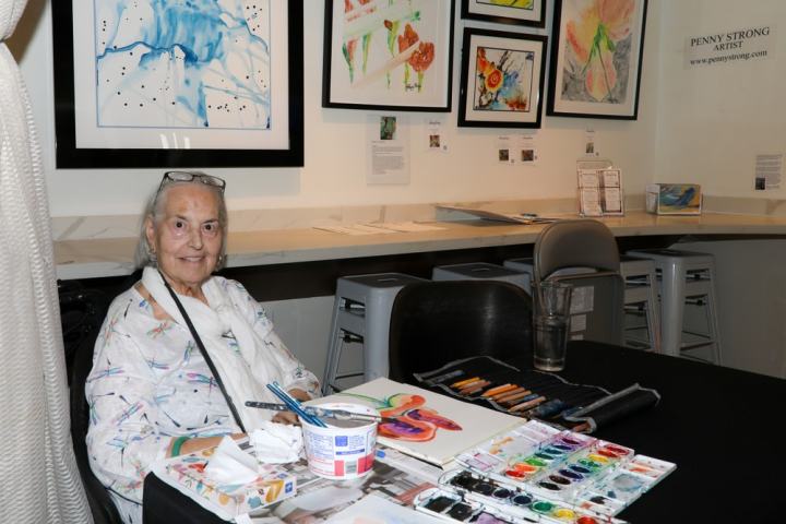 Woman seated at a table with art supplies, paintings on the wall behind her.