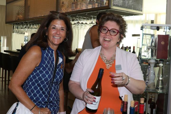 Two women smiling; one holds a wine bottle at a counter with wine corks.