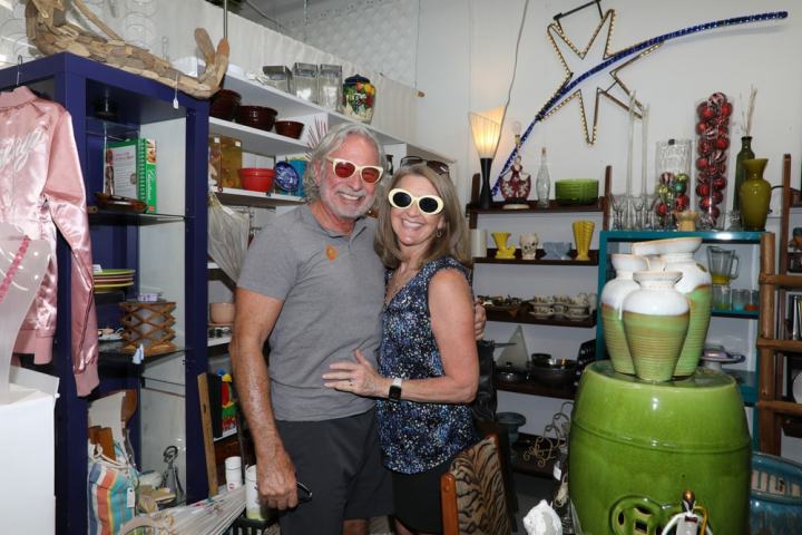 Smiling couple in sunglasses at a vintage shop with shelves of eclectic items.