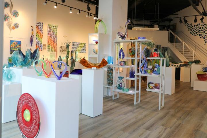 Colorful glass art pieces displayed on white stands in a gallery with wooden floors.