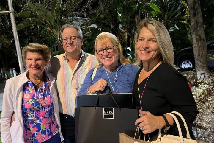 Four smiling adults with shopping bags pose outdoors at night near trees.