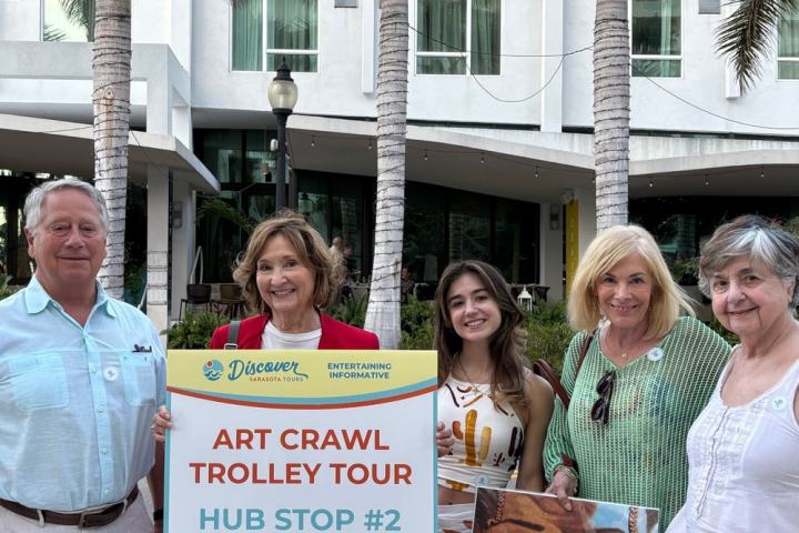 Five people holding an 'Art Crawl Trolley Tour' sign in front of a building with palm trees.