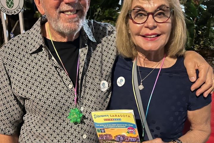 Smiling couple posing outdoors, man with patterned shirt and woman holding a brochure.