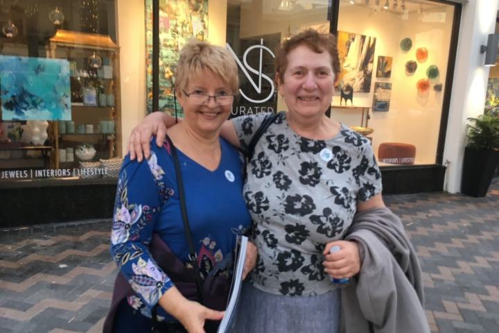 Two smiling women with arms around each other in front of an art gallery window.