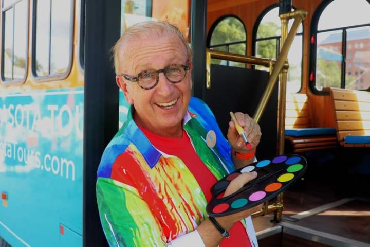 Smiling man in colorful shirt holding a paint palette near a vibrant tour bus.