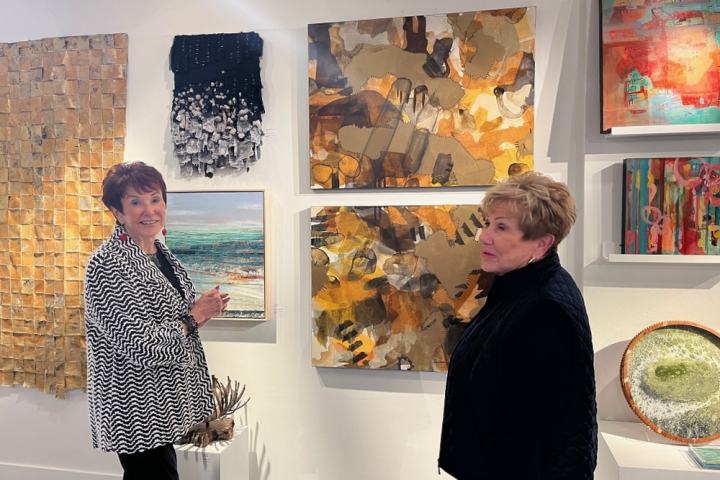 Two women stand in an art gallery with various abstract paintings on the wall.