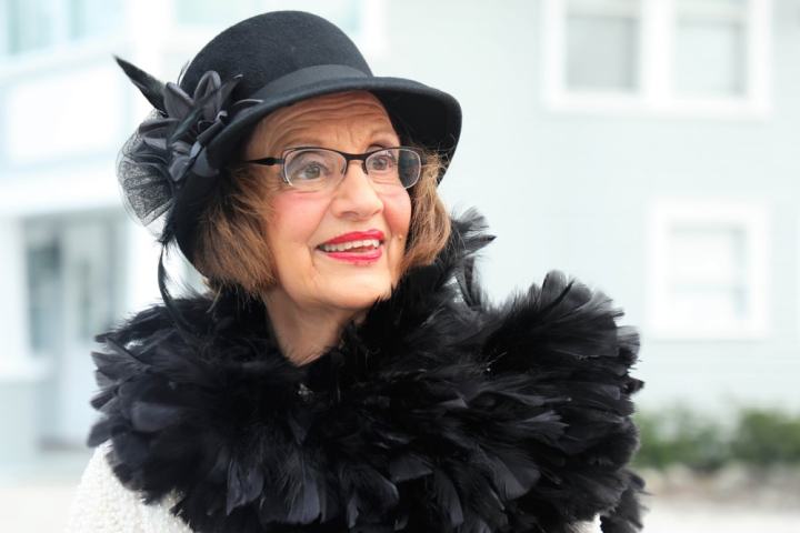 Smiling woman in black hat and feathered scarf outside a building.
