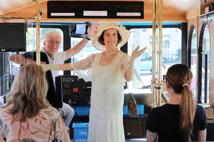 Woman in vintage attire stands in trolley, facing seated passengers, with a driver in the background.