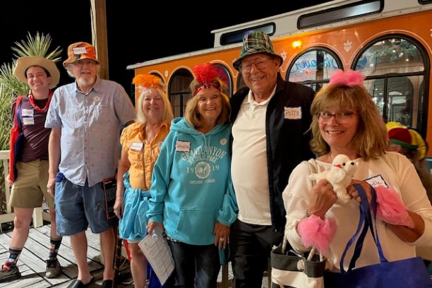 Group of smiling people in colorful outfits and hats standing in front of an orange bus at night.