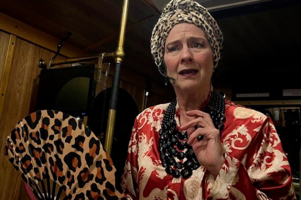 Person in patterned turban and robe holds a leopard-print fan, standing in a dimly lit room.
