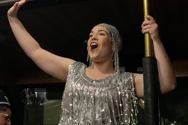 Person in a sparkling dress and headpiece singing with arm raised, holding a pole.