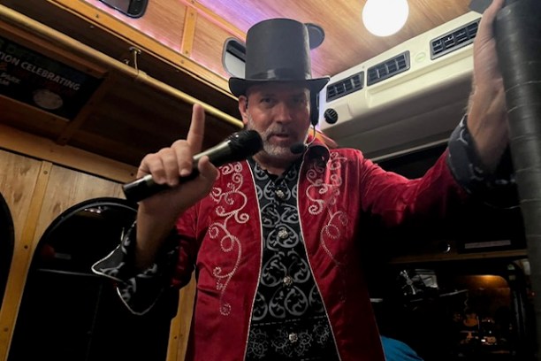 Man in red jacket and top hat holding microphone on a bus.