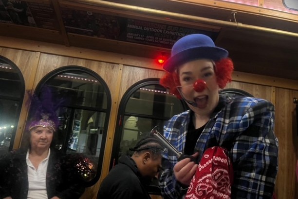 Clown with red nose and blue hat on a bus; people in costumes around.