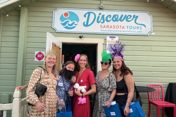 Five women in colorful attire pose with gift bags in front of 'Discover Sarasota Tours' building.