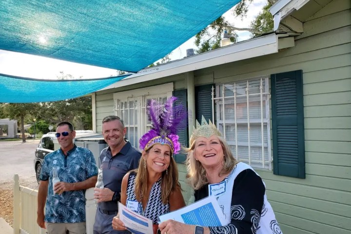Four people smiling outdoors, two wearing decorative hats, holding papers by a green house.