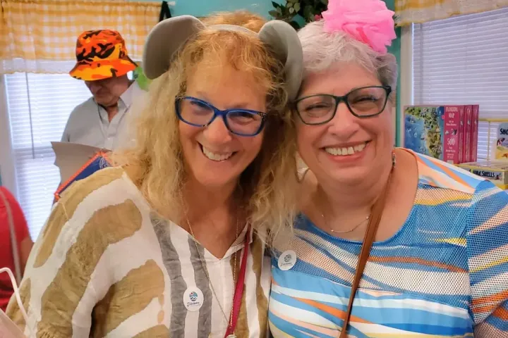Two women smiling, one with horns and the other with a pink bow, inside a room with yellow curtains.