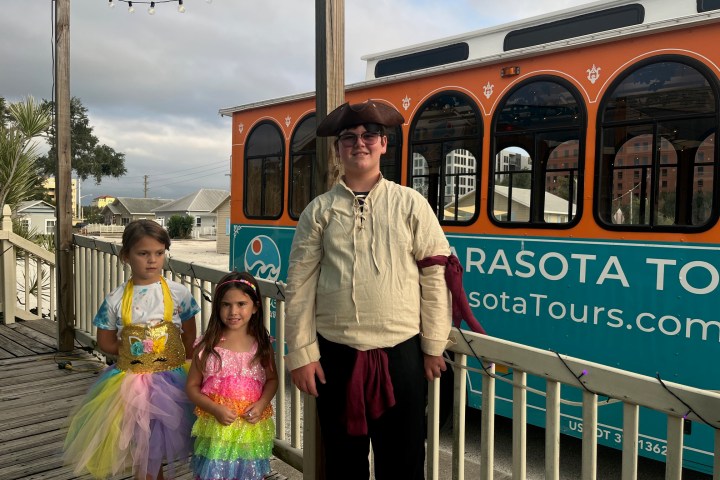 Three children in colorful costumes in front of an orange trolley with 'Sarasota Tours' text.