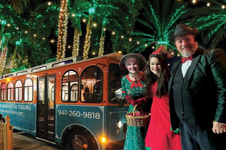 Three people in festive clothes smiling by a brightly lit trolley at night.