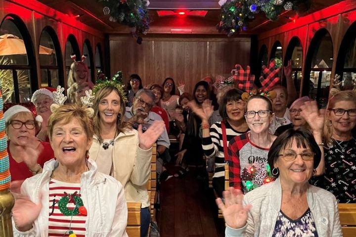 Group of people wearing festive attire and antlers, smiling and waving inside a decorated trolley.