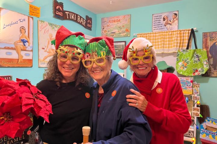 Three women in festive hats and glasses smile in a room with tropical decor.