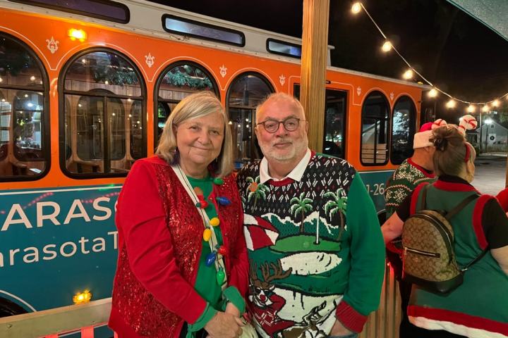 Two people in festive sweaters smiling in front of a decorated trolley at night.