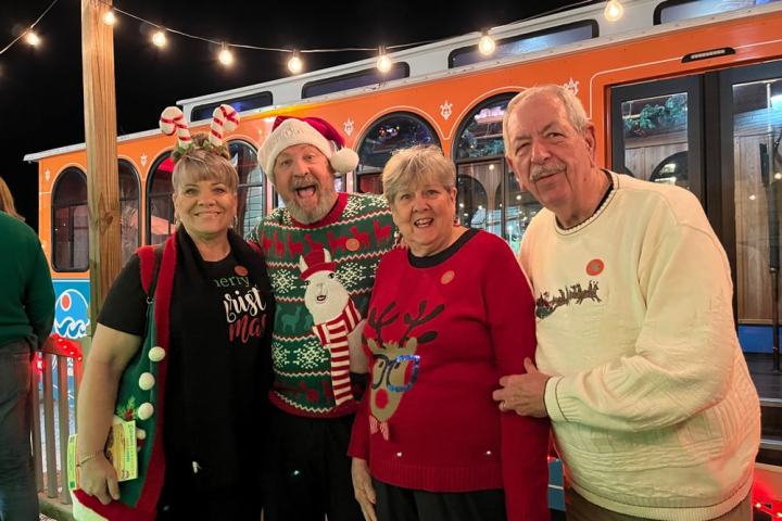 Four people in festive sweaters posing by a decorated trolley at night.