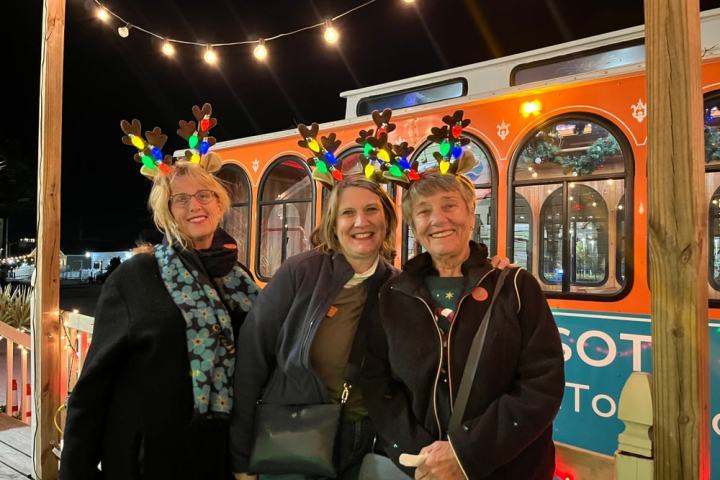 Three women wearing light-up antlers, smiling in front of a decorated trolley at night.
