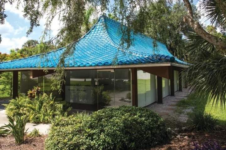 Modern building with blue-tiled roof, glass walls, surrounded by lush greenery.