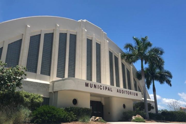White municipal auditorium with arched windows and palm trees under a clear blue sky.