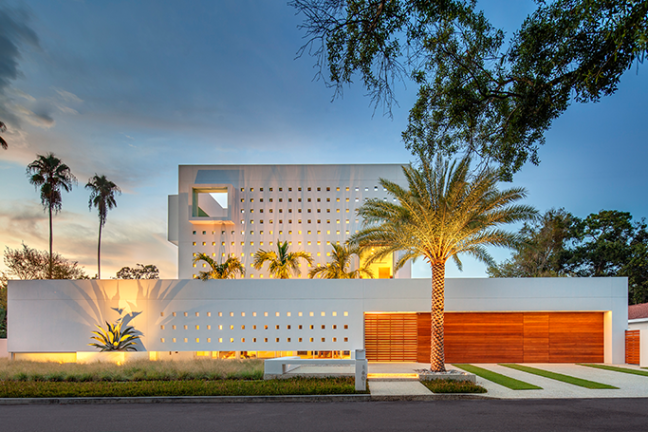 Modern white building with palm trees at sunset, featuring perforated wall design and wooden garage door.
