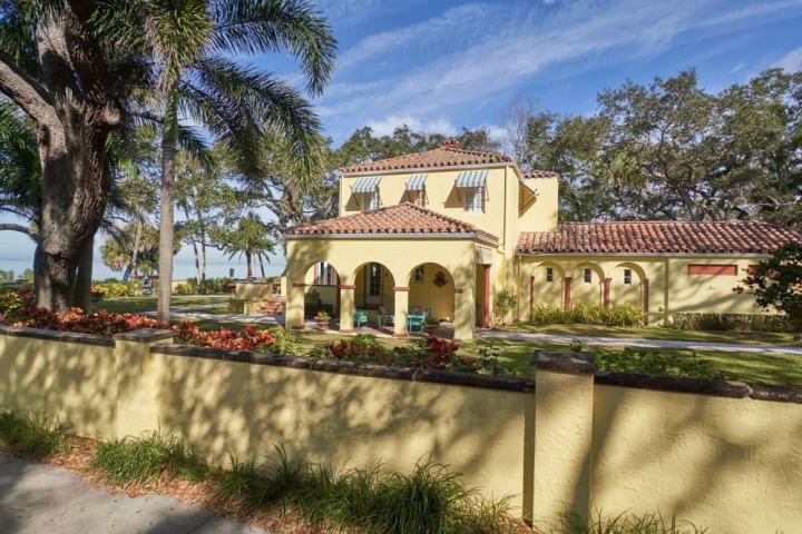 Yellow stucco house with red-tiled roof, surrounded by a garden and palm trees.