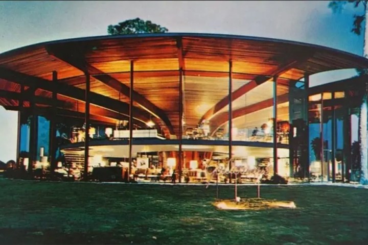 Modern round glass house with warm lighting at night, surrounded by lawn.