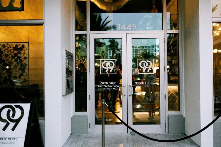 Entrance of bar with '99 Bottles' logo and glass doors, reflecting palm trees and people inside.